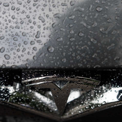 Close-up of a Tesla logo with rain droplets on the surface, highlighting vehicle protection.