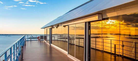 Modern balcony overlooking ocean with sunset reflections on large glass windows.