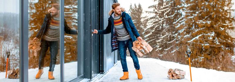 Man in winter attire carrying firewood near a modern cabin surrounded by snow and trees.